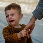 Jak pomóc dziecku radzić sobie z porażką: gry, język i nastawienie Zapłakany maluch w swetrze trzyma dłoń dorosłego na dworze